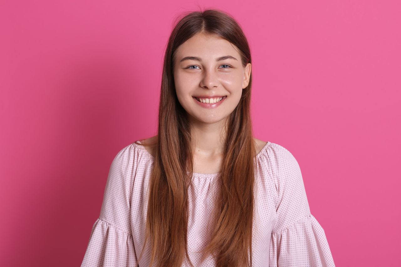 Jeune femme souriante aux cheveux châtains, blouse fluide, portrait doux sur fond rose poudré